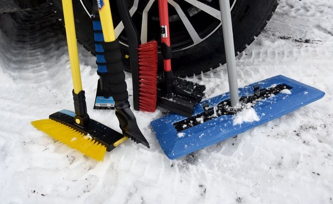 Six different style of snow brush, a snow broom, and ice scrapers on the snow, in front of an Axis wheel with a Bridgestone Blizzak WS90 onit